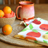 Close up of the DUNS Sweden organic cotton linen tea towel in the citrus print on a wooden table next to a bowl of toy oranges and a red mug