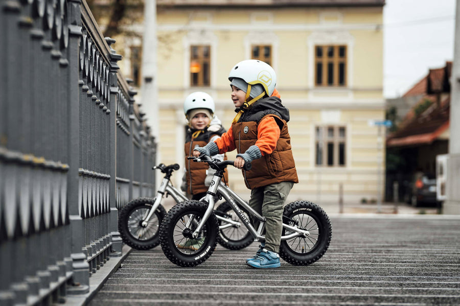 2 children sat on Early Rider big foot balance bikes next to a metal fence