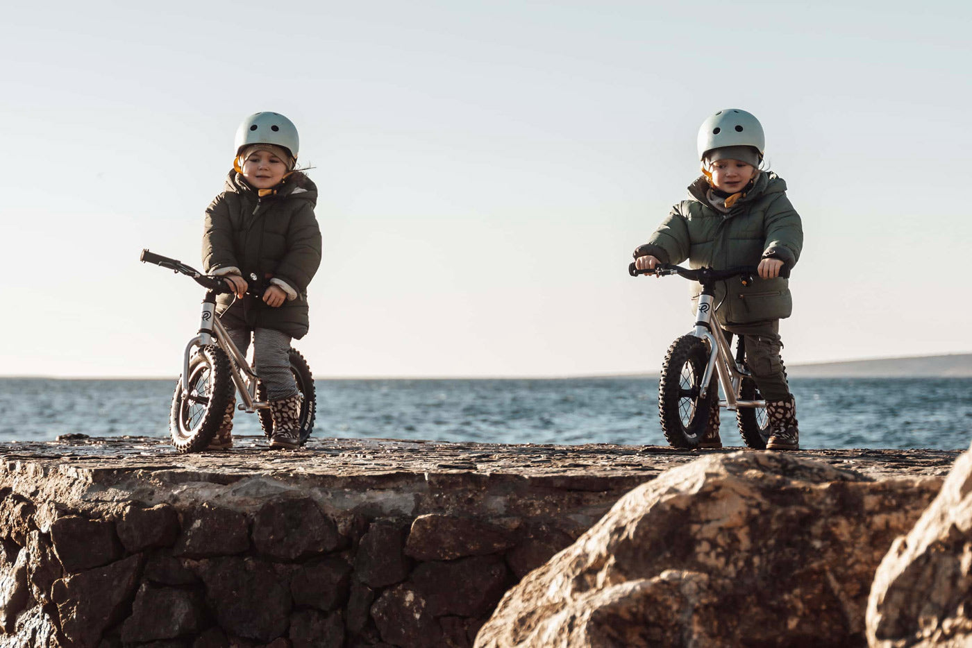 2 children sat on Early Rider aluminium balance bikes on a rock in front of the sea