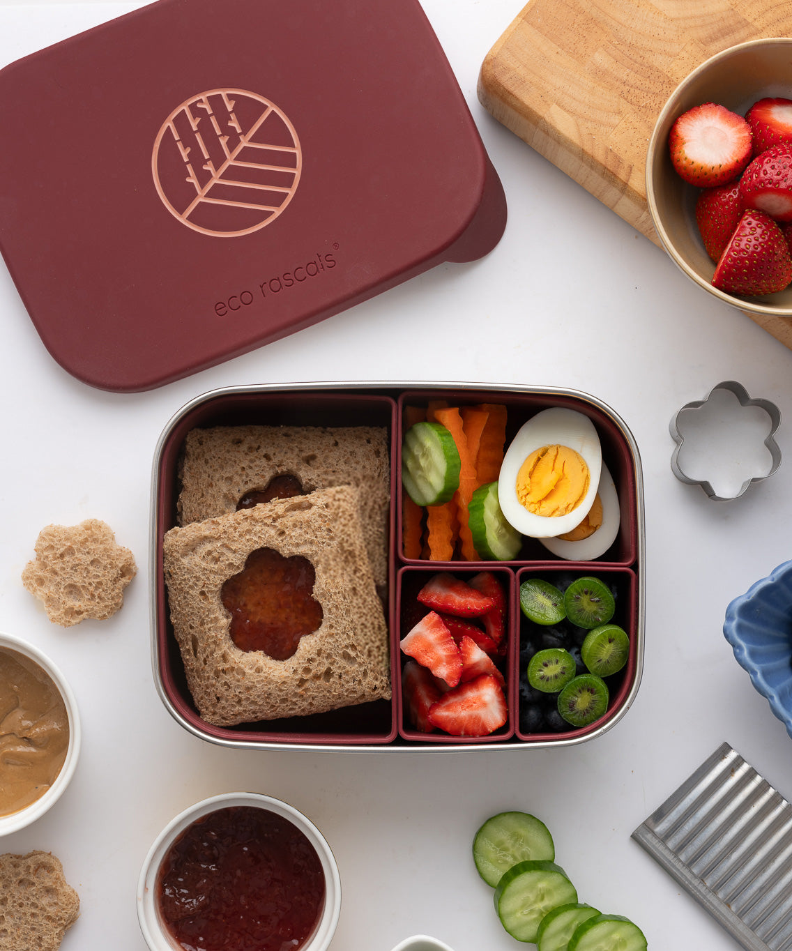 An overhead view of the Eco Rascals Stainless Steel Lunch Box with a burgundy silicone lid and   pot inserts. The lid has been taken off and food can be seen in the lunchbox. As sandwich in the largest section with vegetables, fruit and eggs in the smaller section. 