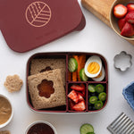 An overhead view of the Eco Rascals Stainless Steel Lunch Box with a burgundy silicone lid and   pot inserts. The lid has been taken off and food can be seen in the lunchbox. As sandwich in the largest section with vegetables, fruit and eggs in the smaller section. 