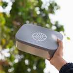 An adult's hand holding up an Eco Rascals Stainless Steel Lunch Box with a silver grey silicone lid.