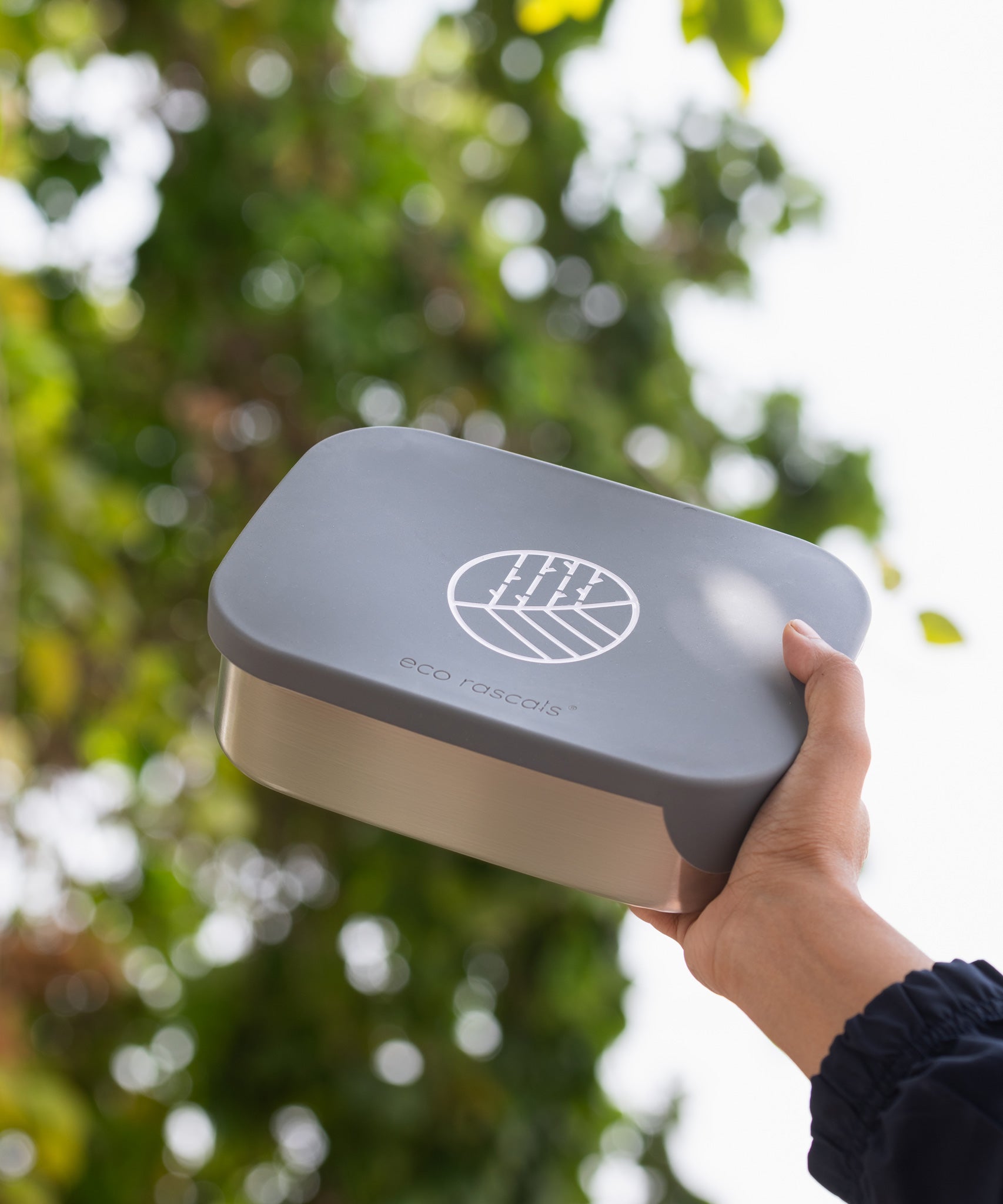 An adult's hand holding up an Eco Rascals Stainless Steel Lunch Box with a silver grey silicone lid.