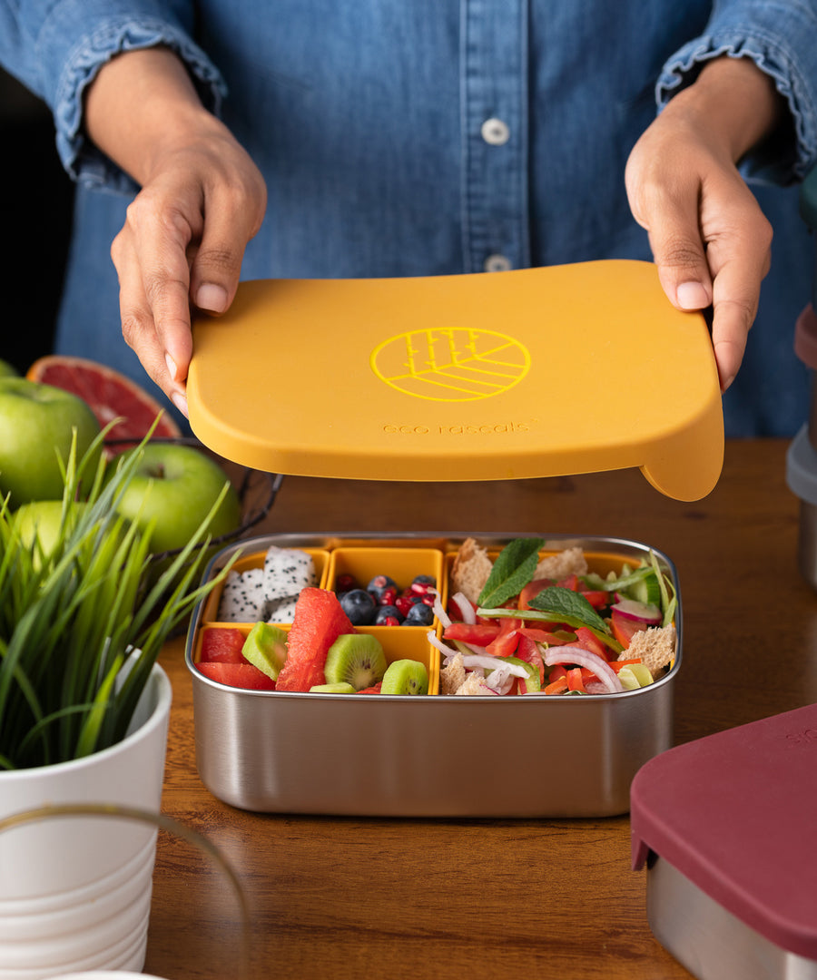 An adults hands lifting the lid off a Eco Rascals Stainless Steel Lunch Box with a mustard yellow silicone lid and pot inserts. The lunchbox is filled with food.