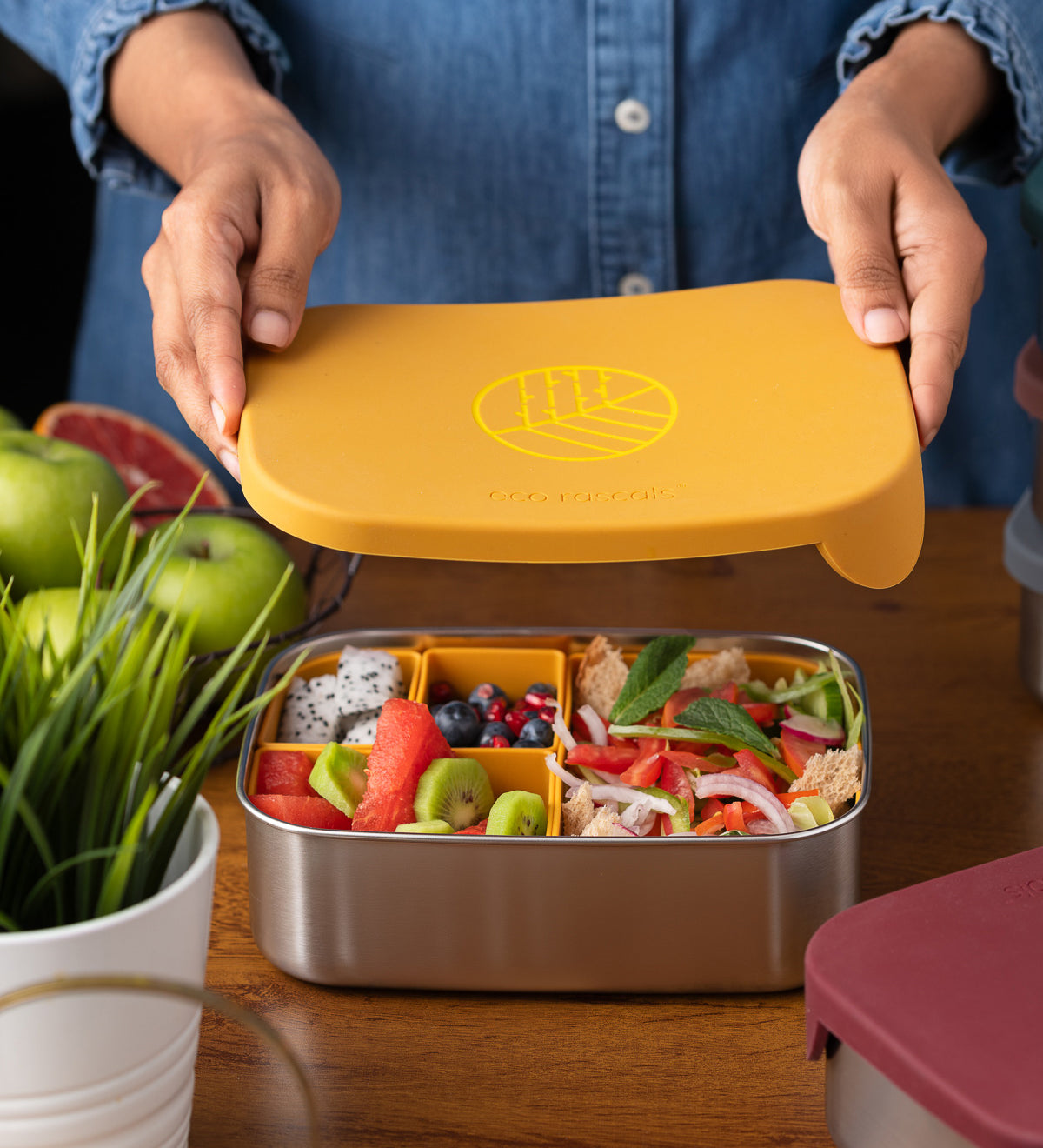 An adults hands lifting the lid off a Eco Rascals Stainless Steel Lunch Box with a mustard yellow silicone lid and pot inserts. The lunchbox is filled with food.