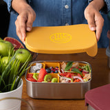 An adults hands lifting the lid off a Eco Rascals Stainless Steel Lunch Box with a mustard yellow silicone lid and pot inserts. The lunchbox is filled with food.