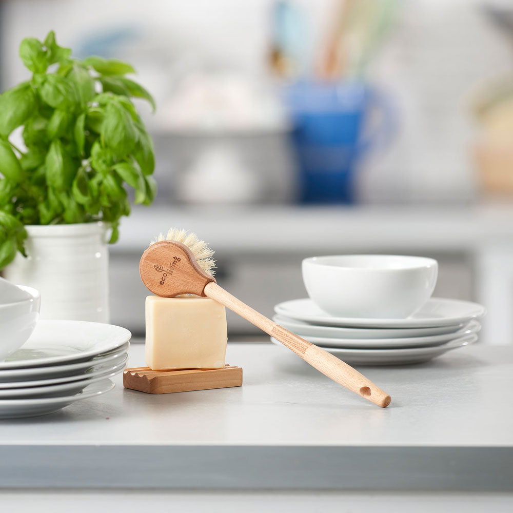 Ecoliving Long Handle Wooden Dish Brush pictured on a dish soap bar resting on a wooden soap dish with stacks of white ceramic bowls and plates in the background