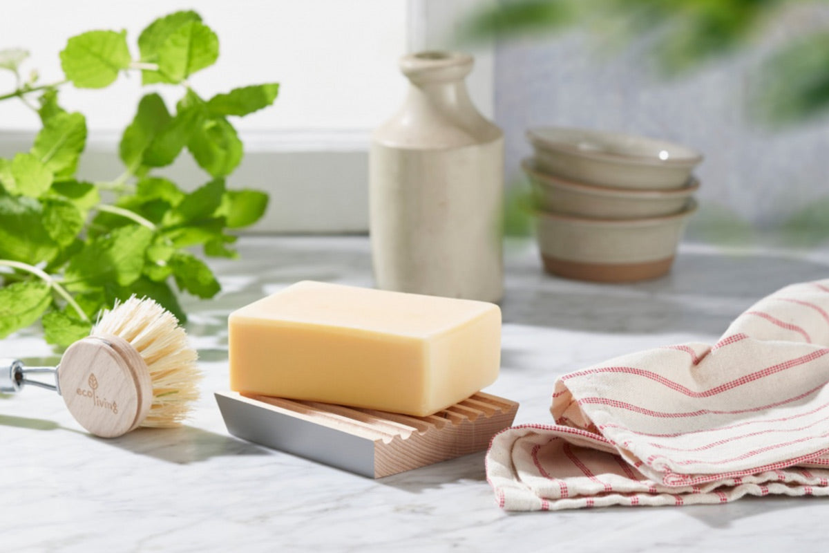 Ecoliving beech wood soap dish with a cream soap on top on a grey marble work top next to a tea towel and a green plant