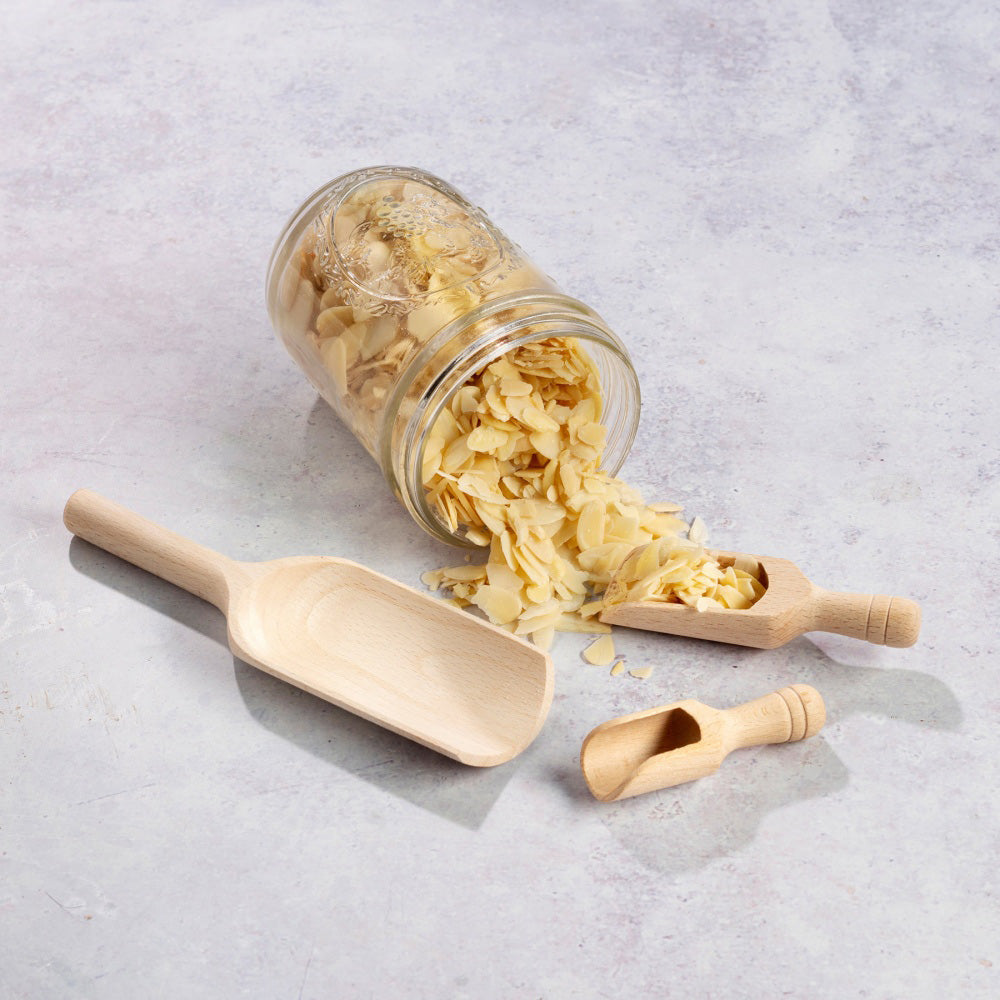 3 Ecoliving solid wooden scoops laid out on a grey background next to an overturned jar of nuts