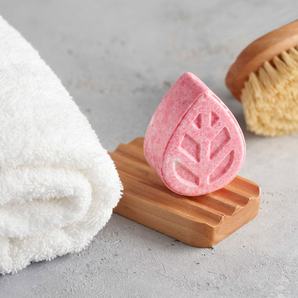 Close up of an Ecoliving solid shampoo sample bar on a wooden soap dish next to a white towel