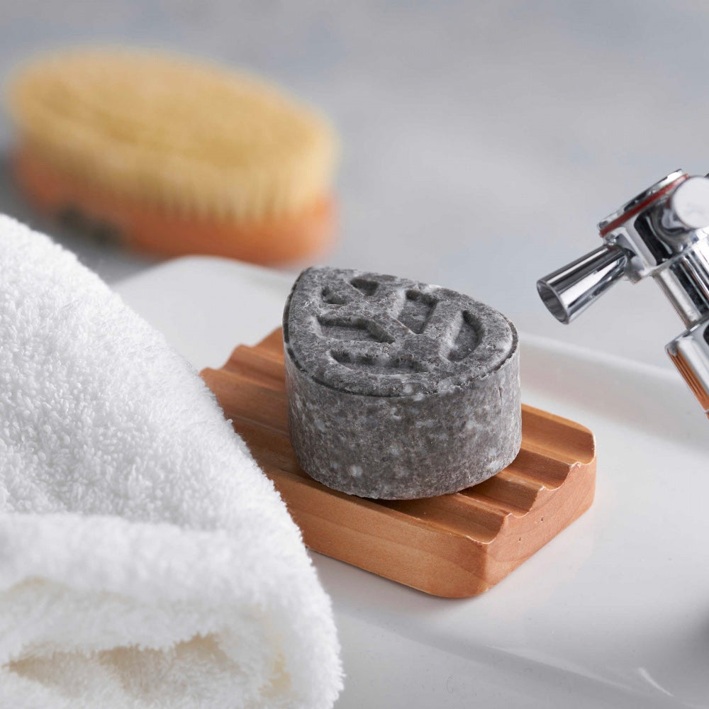 Close up of an Ecoliving grey solid shampoo sample bar on a wooden soap dish next to a white towel