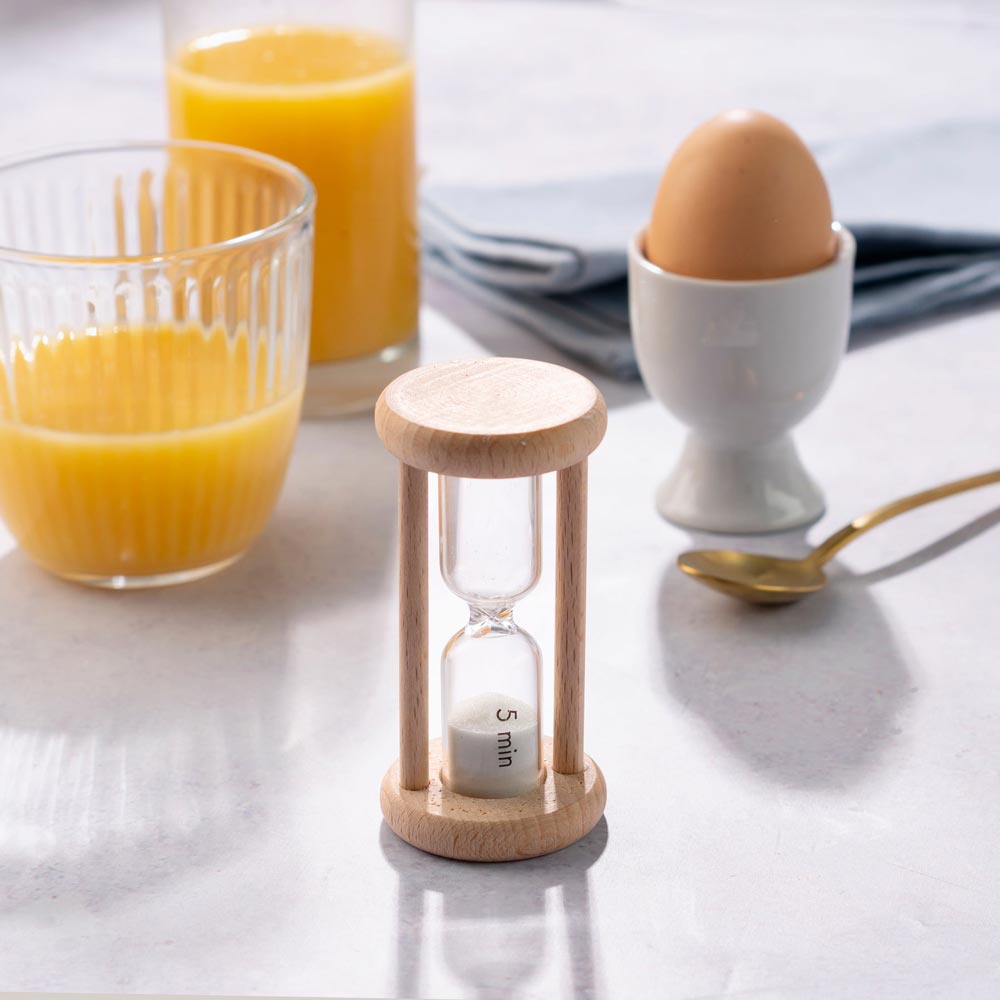 Ecoliving Wooden Egg Timer pictured in front of a boiled egg in a ceramic holder and 2 glasses of orange juice in the background