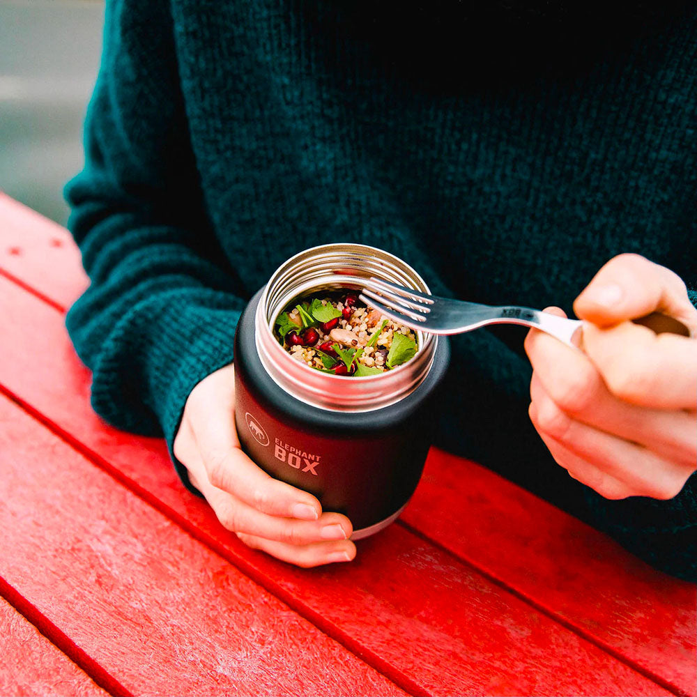 Close up of someone using a fork to eat out of the Elephant Box stainless steel insulated food flask on a red bench