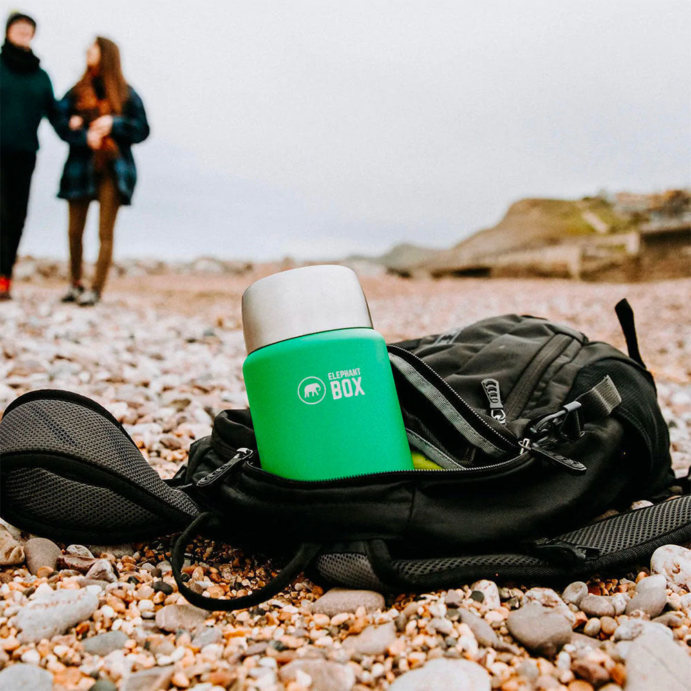 Close up of a green Elephant Box steel food flask in a black backpack on a pebble beach