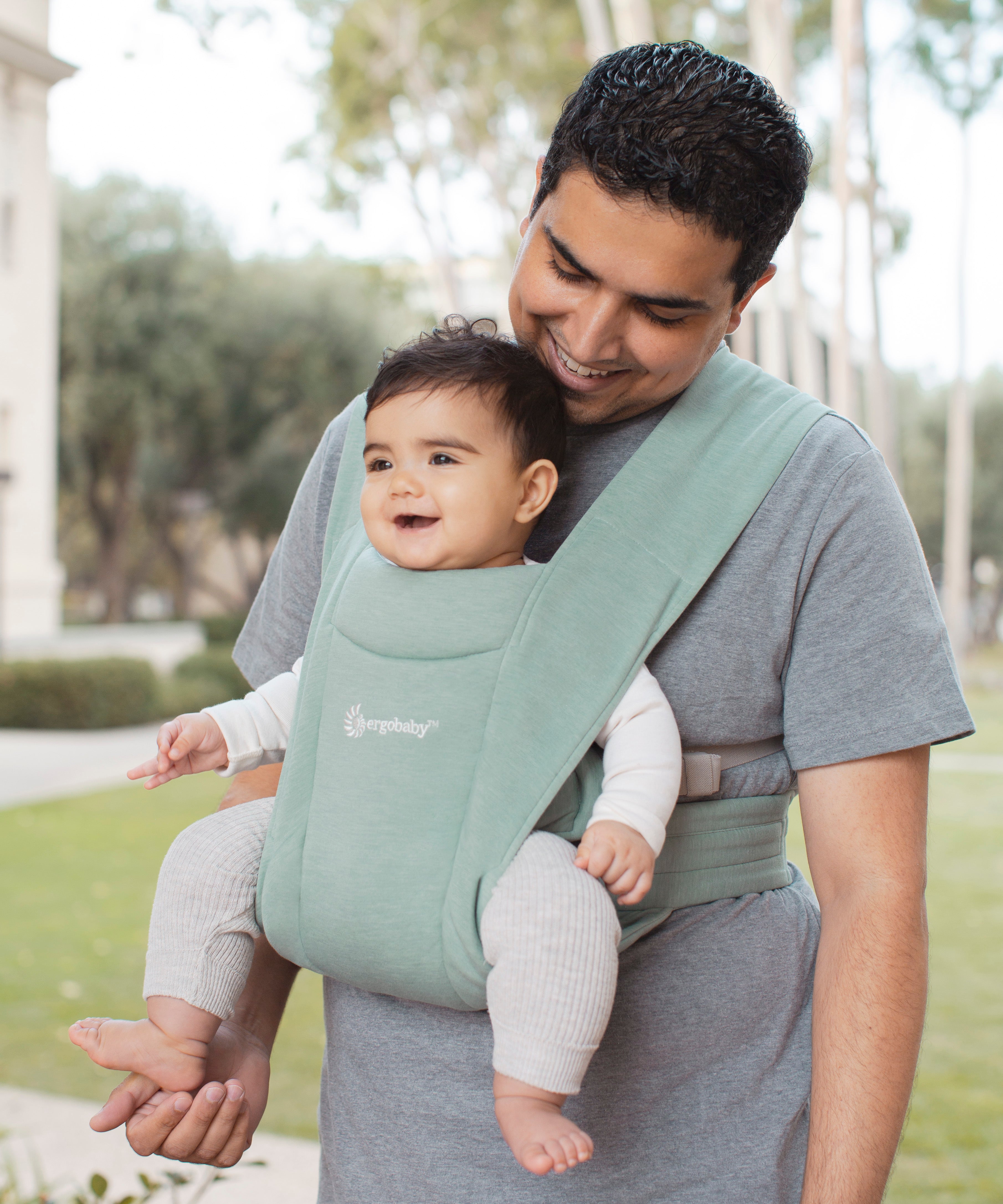 Man wearing a grey t-shirt and his baby on his front facing outwards in the Ergobaby Embrace Soft Knit Newborn Baby Carrier in Jade. Side view in a garden.