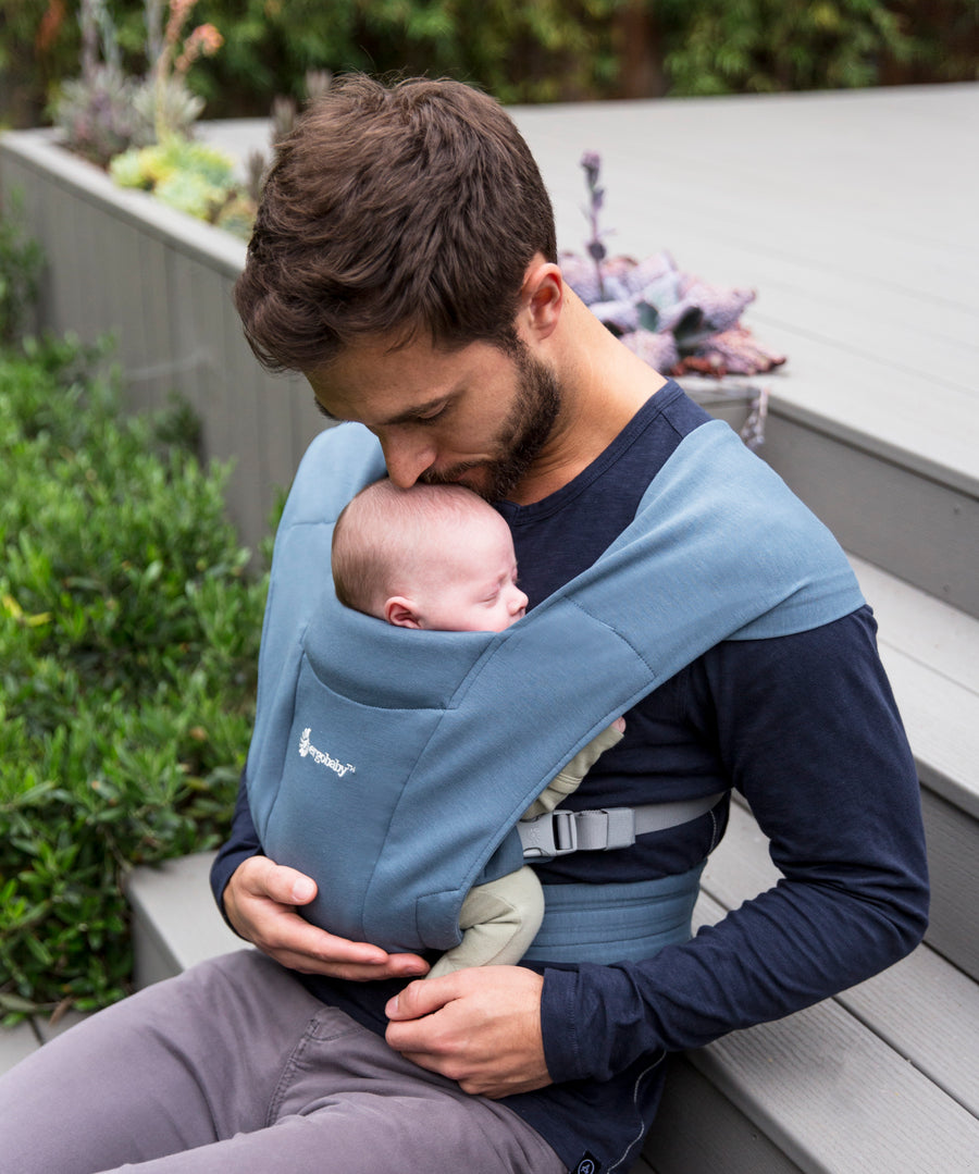 Man wearing a navy blue top and his baby on his front facing inwards in the Ergobaby Embrace Soft Knit Newborn Baby Carrier in Oxford Blue. Side view sat on a garden bench.