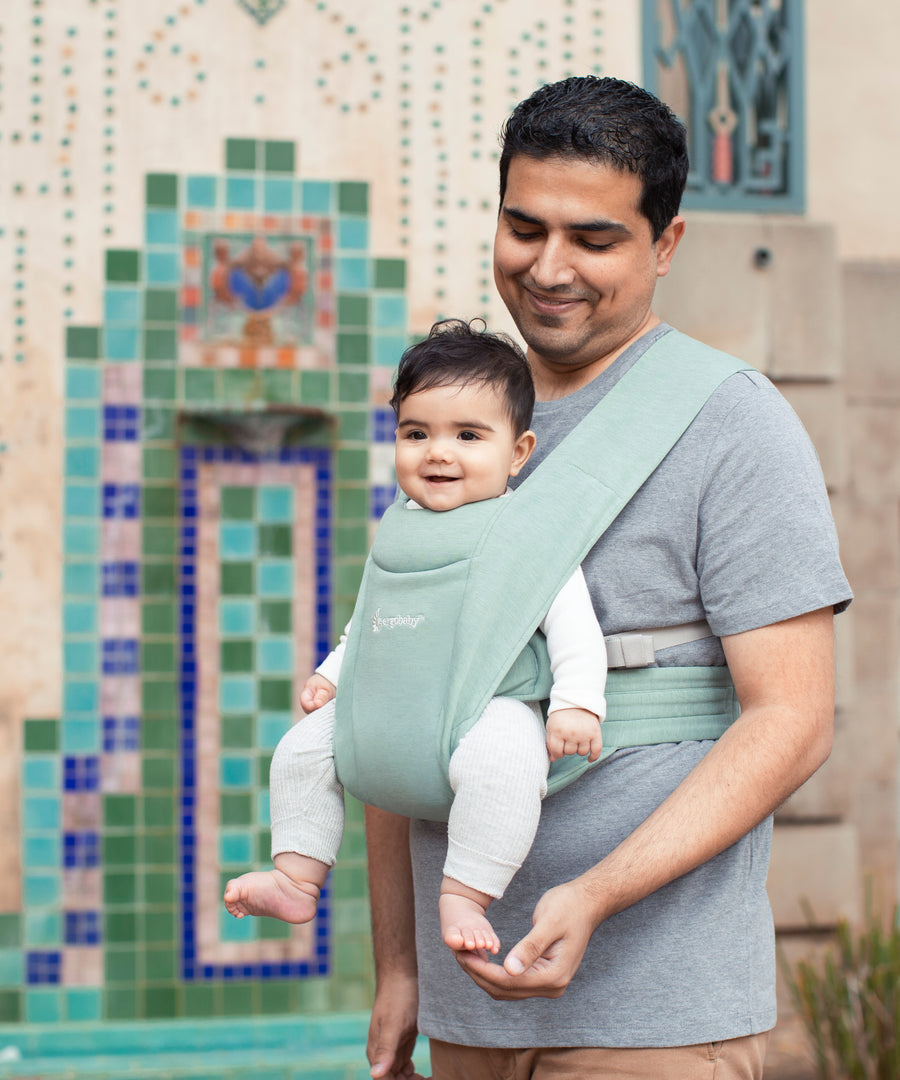 Man wearing a grey t-shirt and his baby on his front facing outwards in the Ergobaby Embrace Soft Knit Newborn Baby Carrier in Jade. Side view in front of a colourful tiled wall.