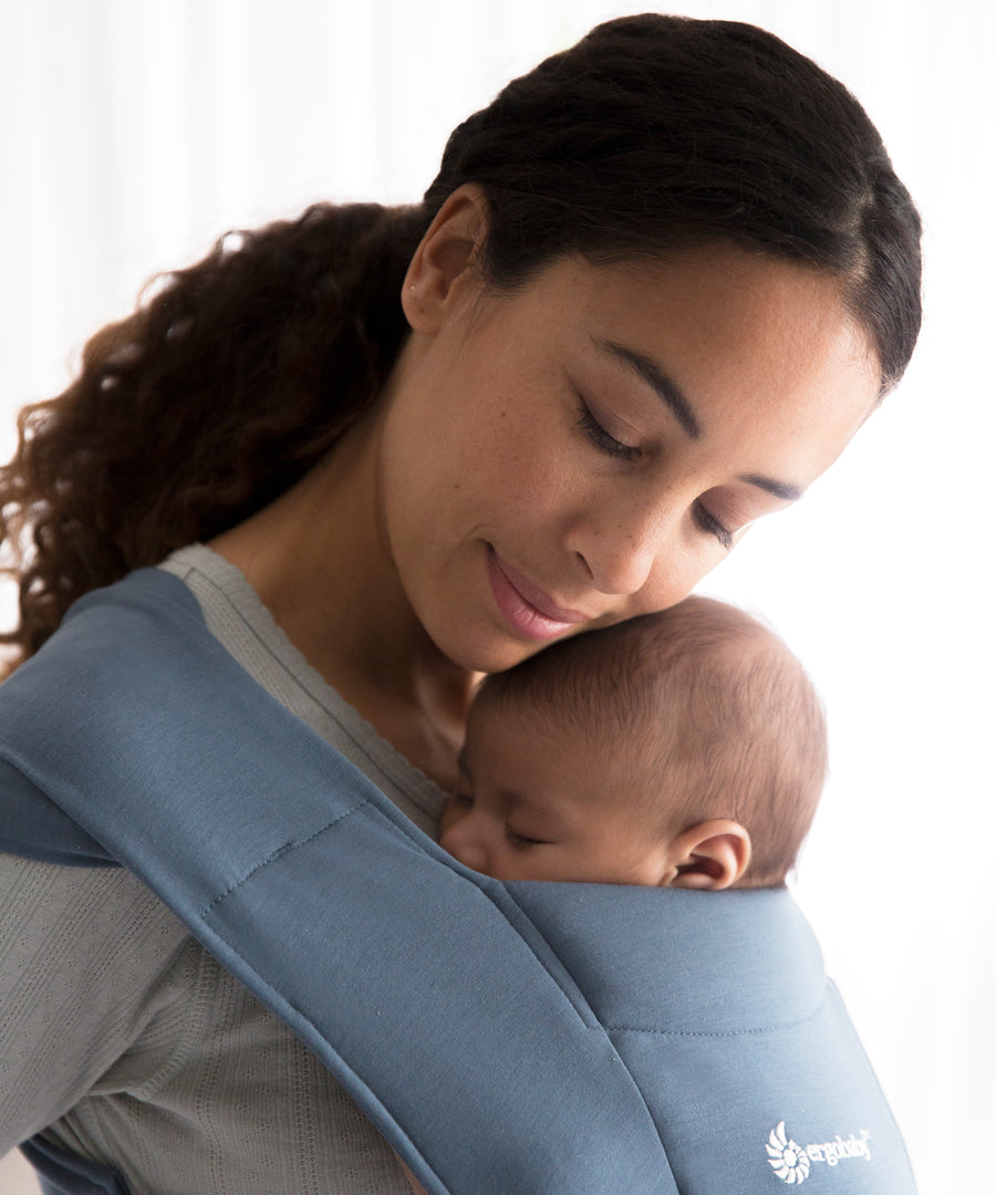 Woman wearing a grey sweater and her sleeping baby on her front in the Ergobaby Embrace Soft Knit Newborn Baby Carrier in Oxford Blue. Side view white background.