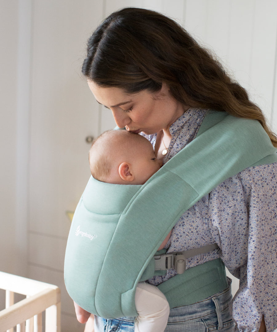 Woman wearing a floral blouse and her baby on her front facing inwards in the Ergobaby Embrace Soft Knit Newborn Baby Carrier in Jade. Side view next to a cot.