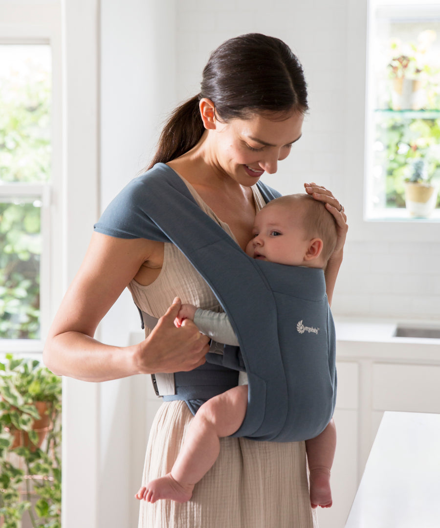 Woman wearing a white vest and her baby on her front facing inwards in the Ergobaby Embrace Soft Knit Newborn Baby Carrier in Oxford Blue. Side view, looking down to stroke baby's head.