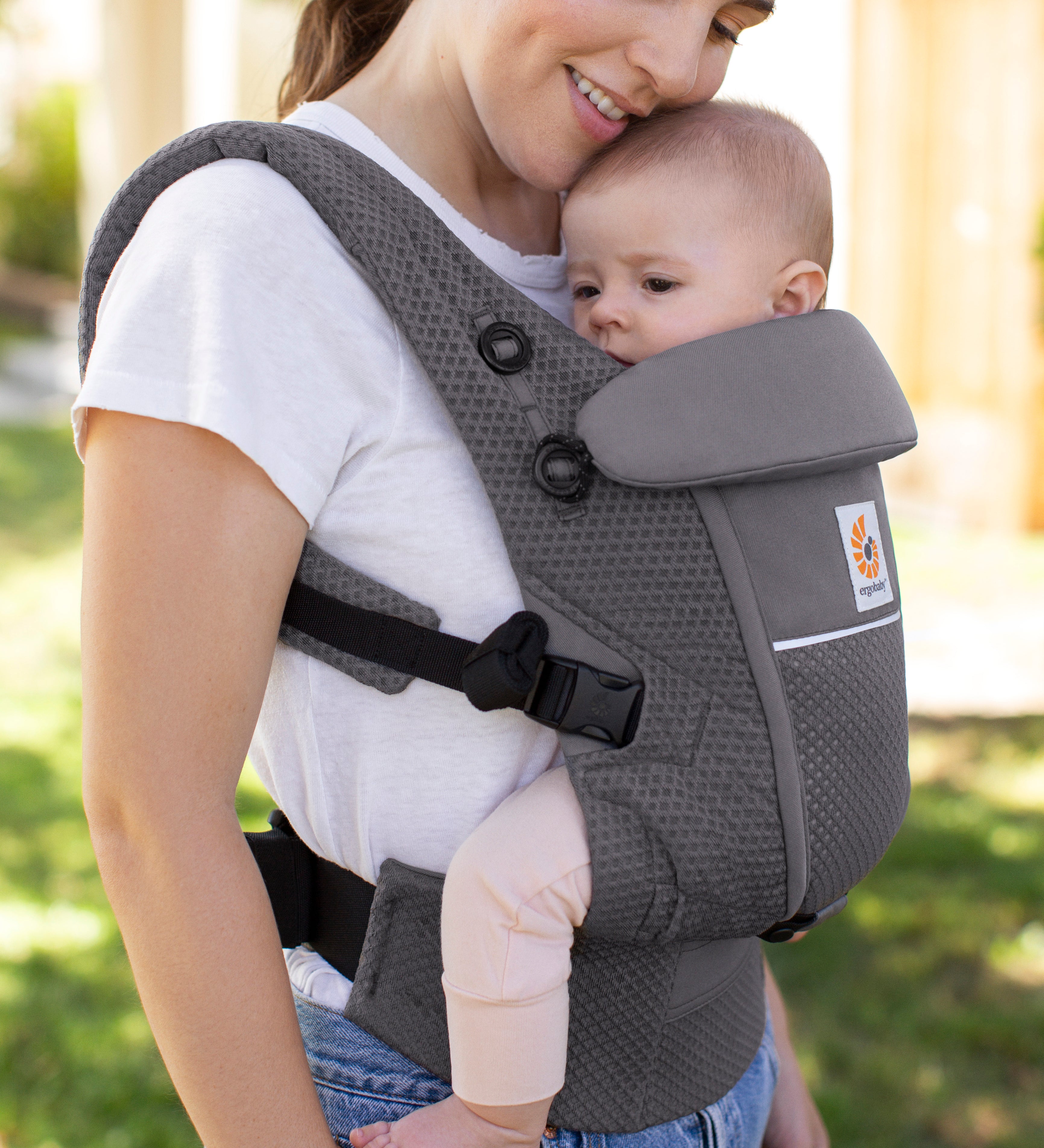 Woman with brown hair and a white t-shirt wearing her baby in the Ergobaby Adapt  SoftFlex™ Mesh Baby Carrier in Graphite Grey. Front view, outside in a garden. 