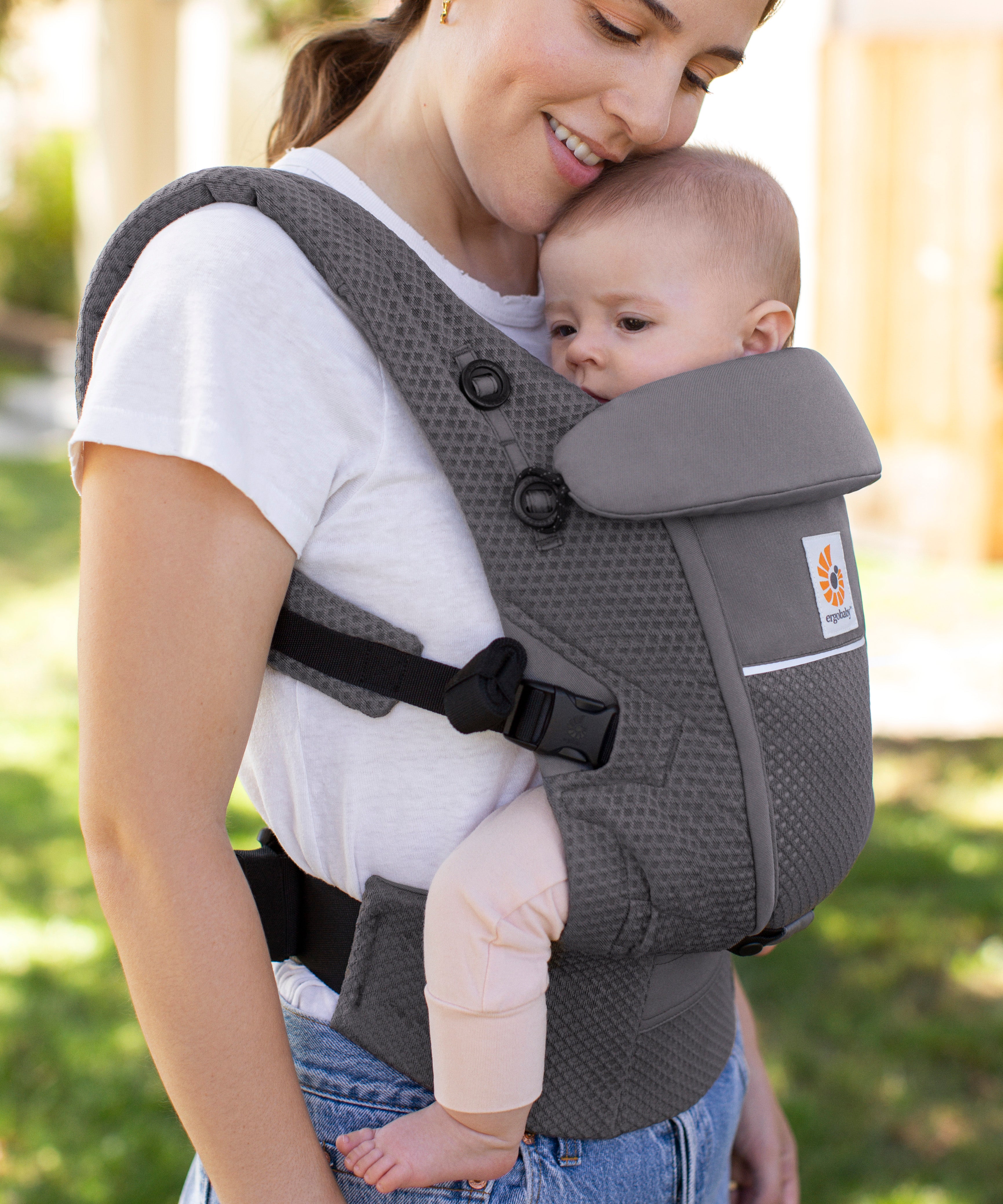 Woman with brown hair and a white t-shirt wearing her baby in the Ergobaby Adapt  SoftFlex™ Mesh Baby Carrier in Graphite Grey. Front view, outside in a garden. 