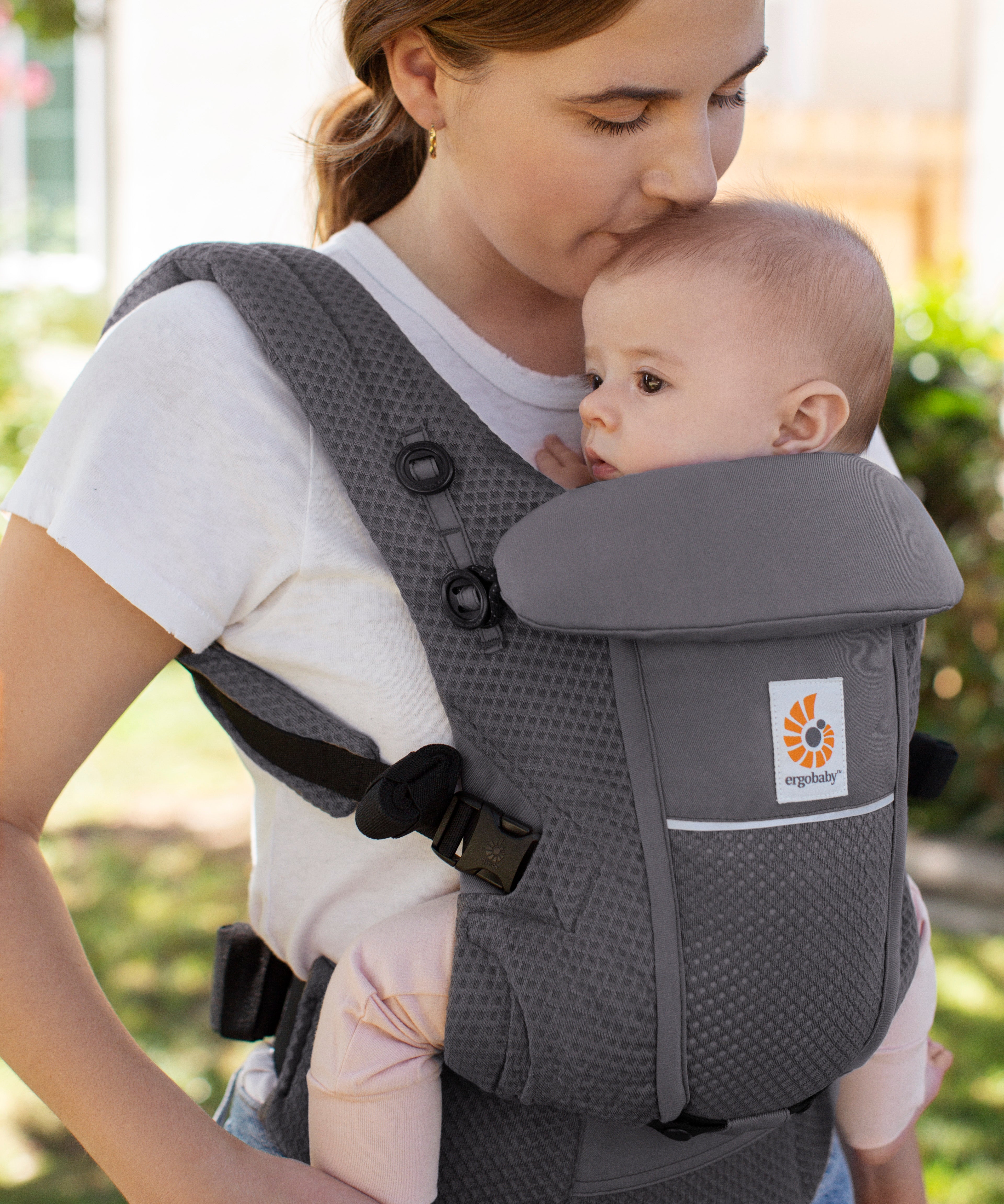 Woman with brown hair and a white t-shirt wearing her baby in the Ergobaby Adapt  SoftFlex™ Mesh Baby Carrier in Graphite Grey. Front view, outside in a garden. 