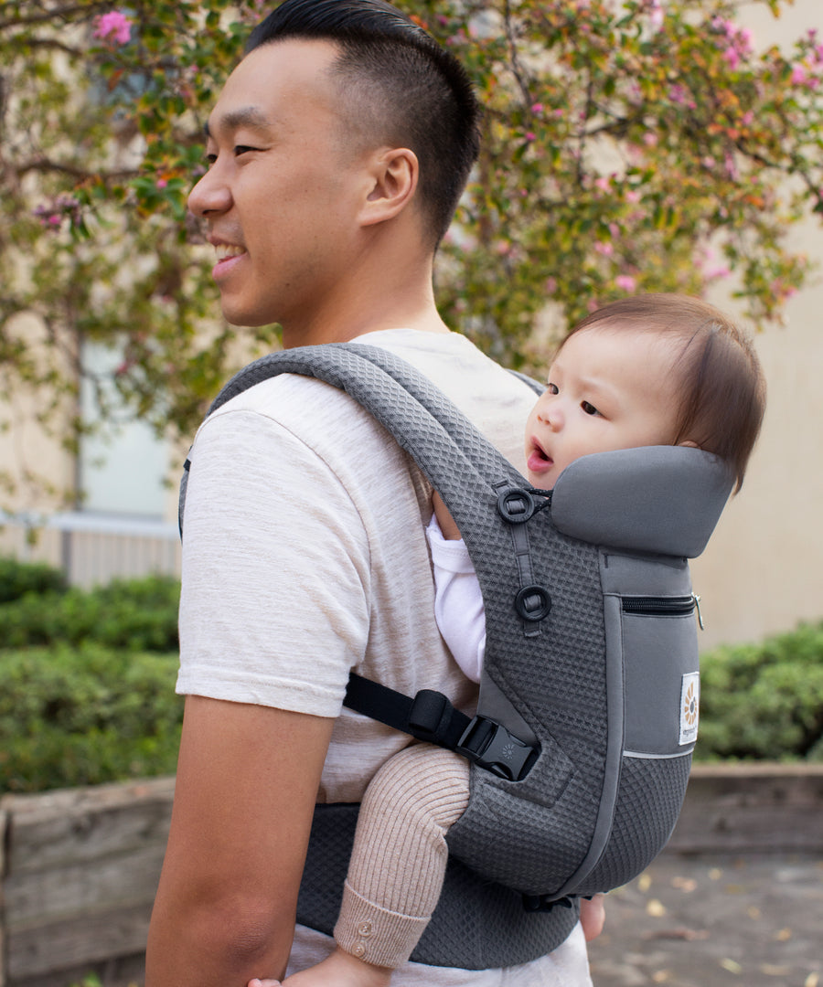 Man with black hair and a white t-shirt wearing his baby on his back in the Ergobaby Adapt  SoftFlex™ Mesh Baby Carrier in Graphite Grey. Side view, outside in a garden. 