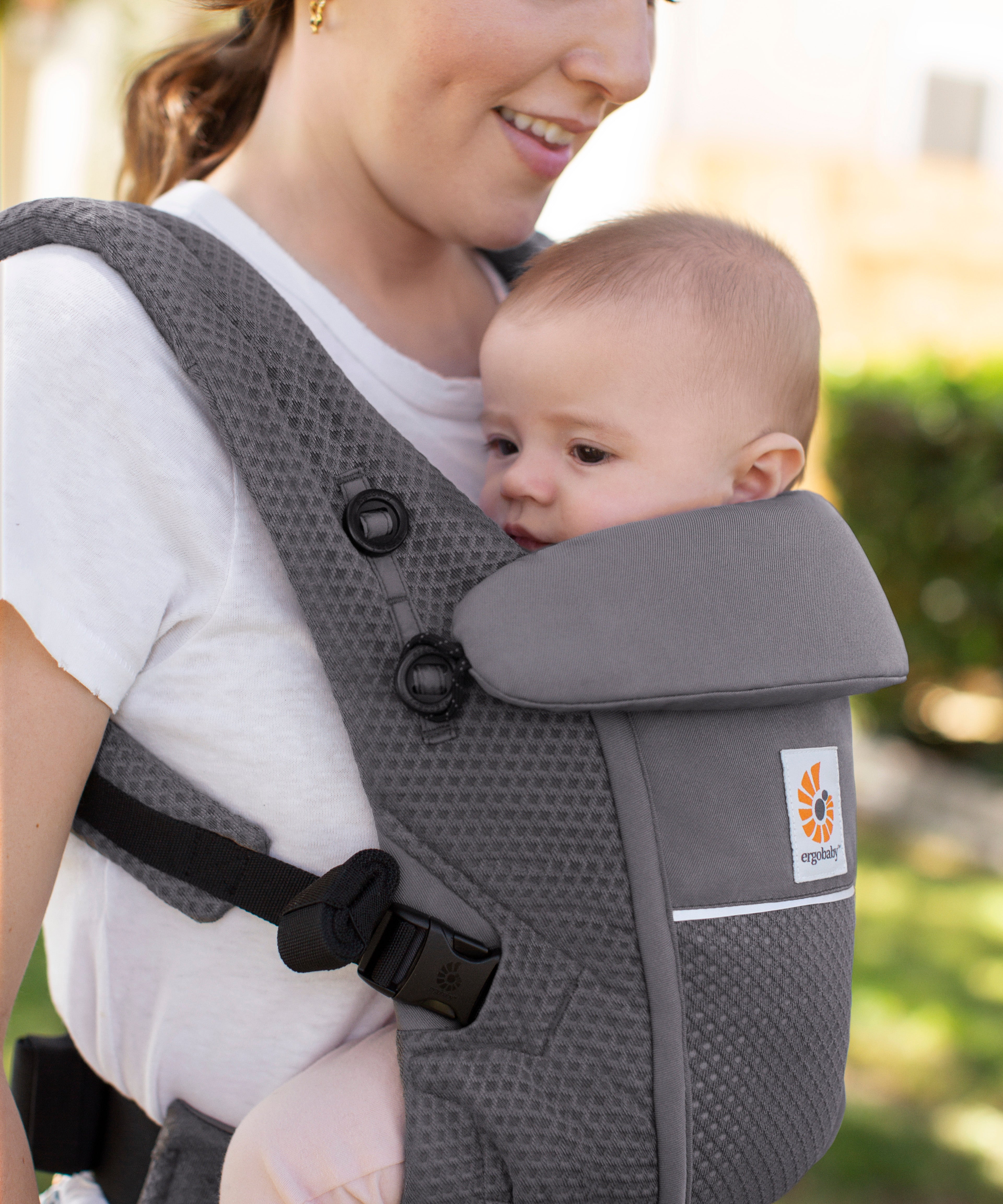 Woman with brown hair and a white t-shirt wearing her baby in the Ergobaby Adapt  SoftFlex™ Mesh Baby Carrier in Graphite Grey. Front view, outside in a garden. 