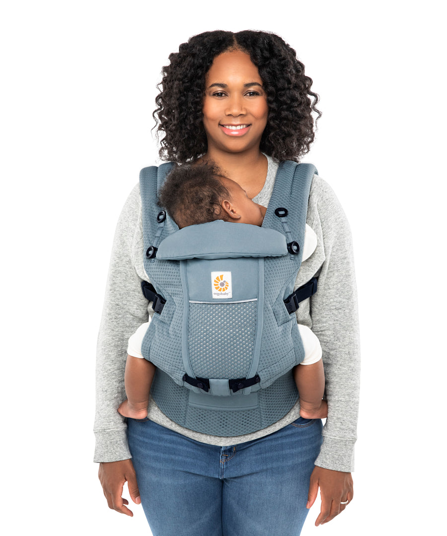 Woman with black curly hair and a grey jumper wearing her baby in the Ergobaby Adapt  SoftFlex™ Mesh Baby Carrier in Slate Blue. Front view, white background. 