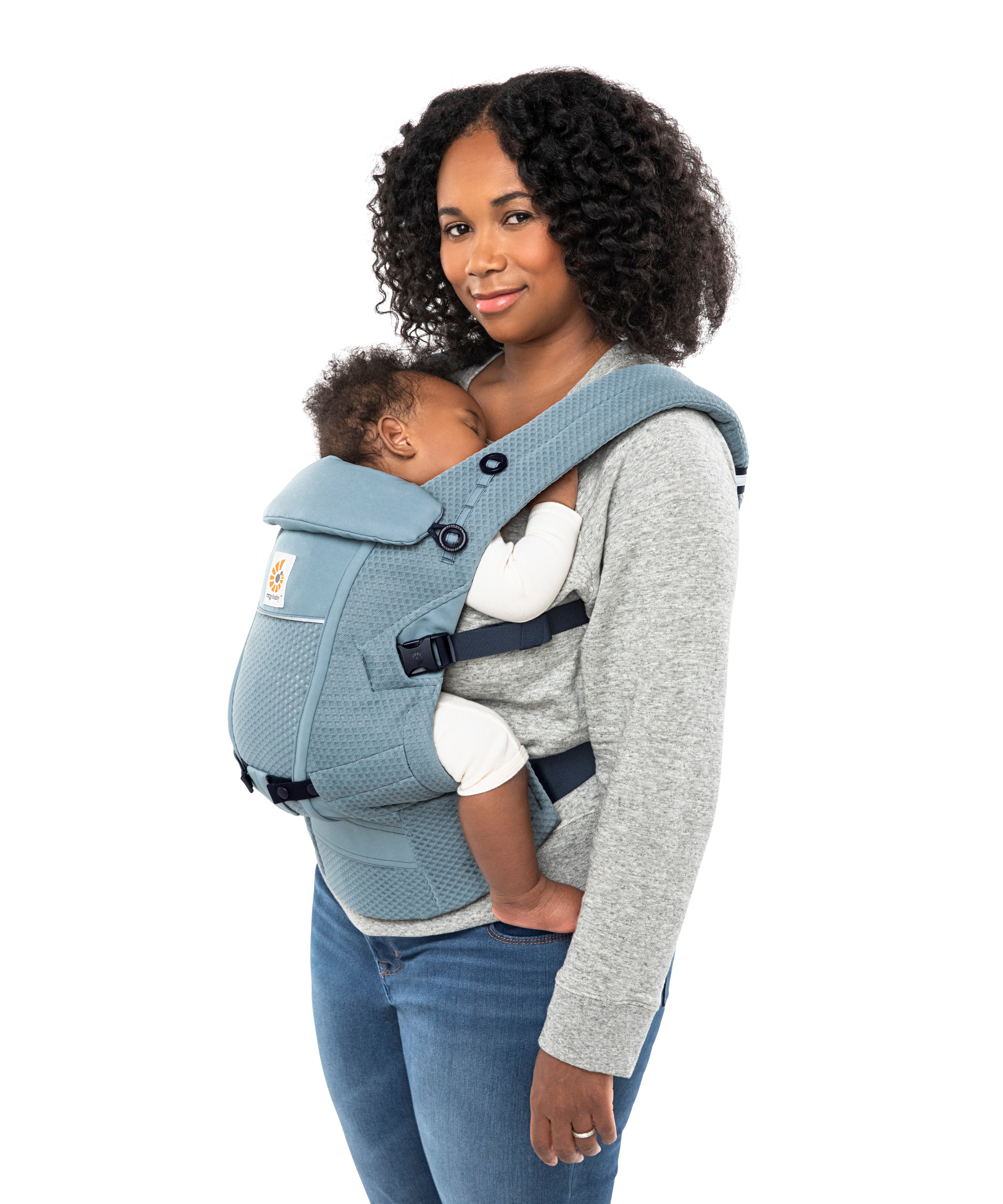 Woman with black curly hair and a grey jumper wearing her baby in the Ergobaby Adapt  SoftFlex™ Mesh Baby Carrier in Slate Blue. Side view, white background. 