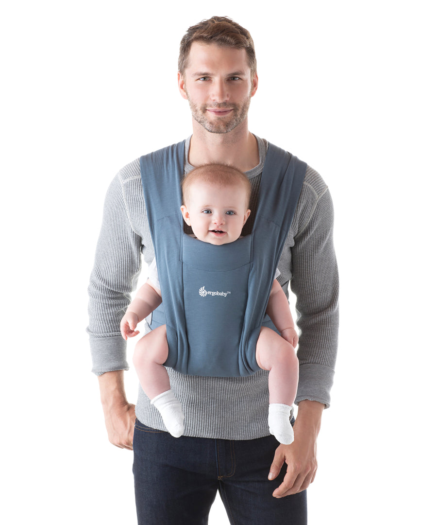 Man wearing a grey jumper and his baby on his front facing outwards in the Ergobaby Embrace Soft Knit Newborn Baby Carrier in Oxford Blue. Front view white background.