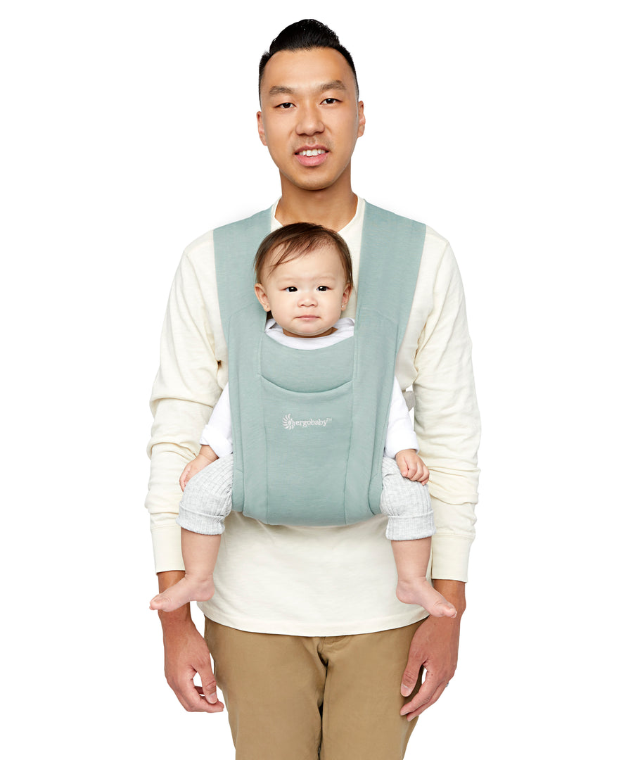 Man wearing a white top and his baby on his front facing outwards in the Ergobaby Embrace Soft Knit Newborn Baby Carrier in Oxford Blue. Front view white background.