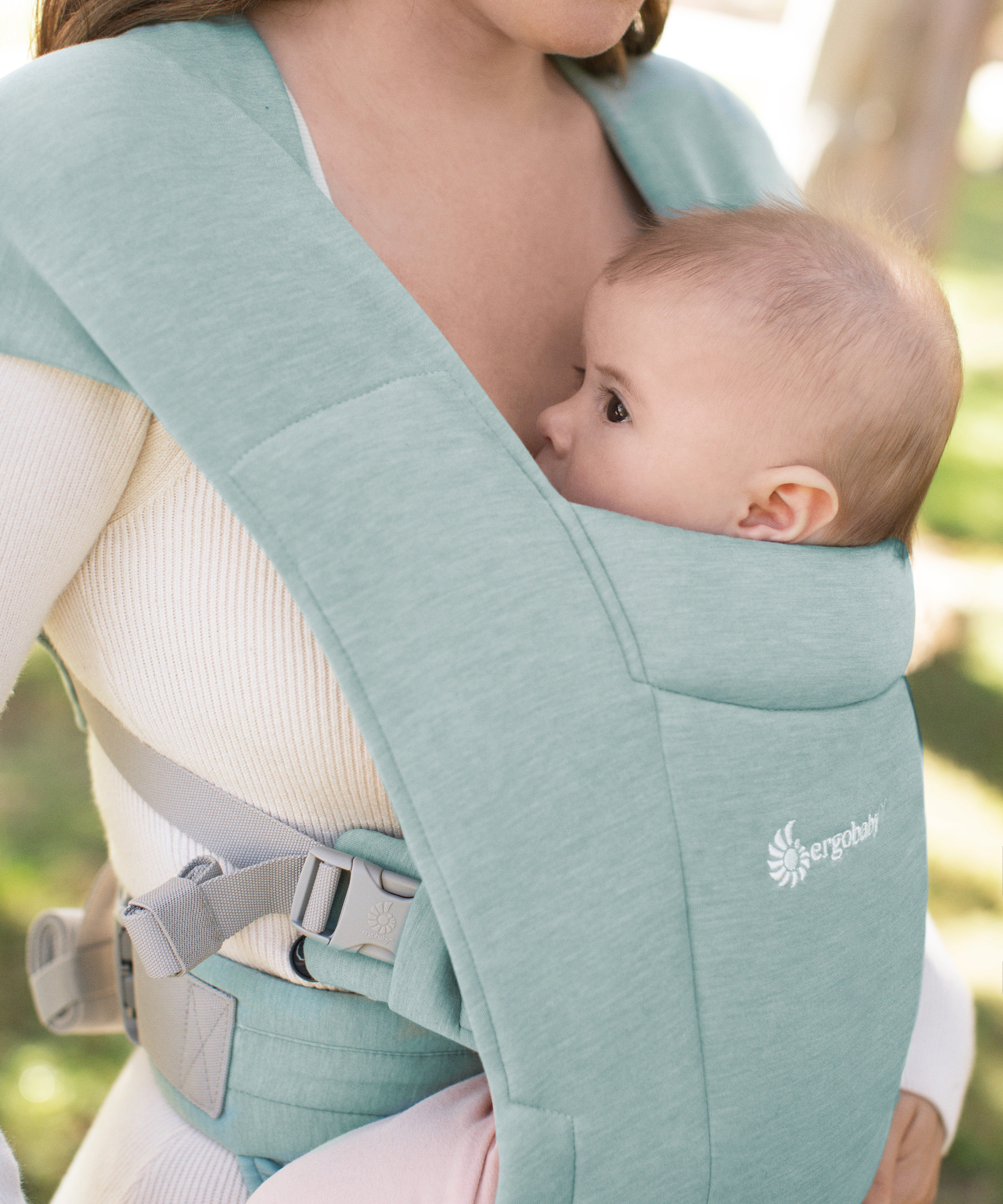 Close up of a baby facing inwards in the Ergobaby Embrace Soft Knit Newborn Baby Carrier in Jade. Side view garden background.