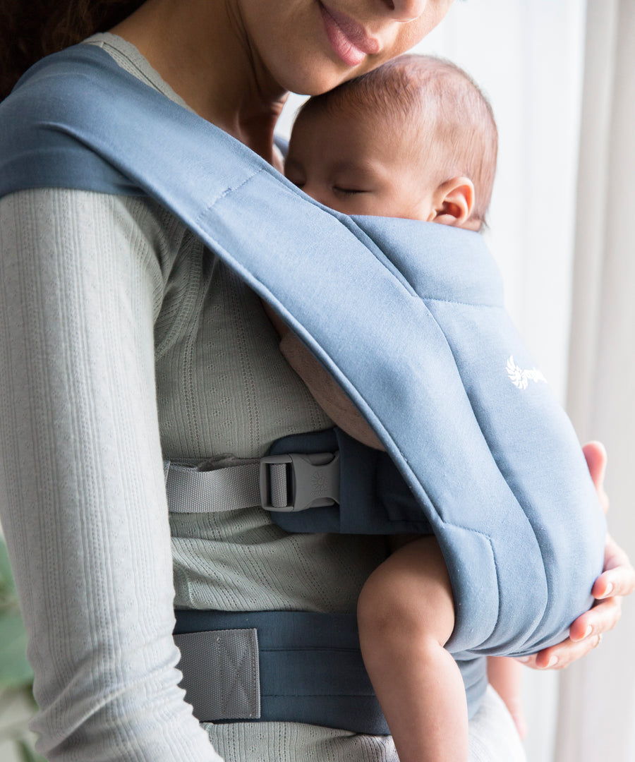 Woman wearing a grey top and her sleeping baby on her front facing inwards in the Ergobaby Embrace Soft Knit Newborn Baby Carrier in Oxford Blue. Side view in front of a window.