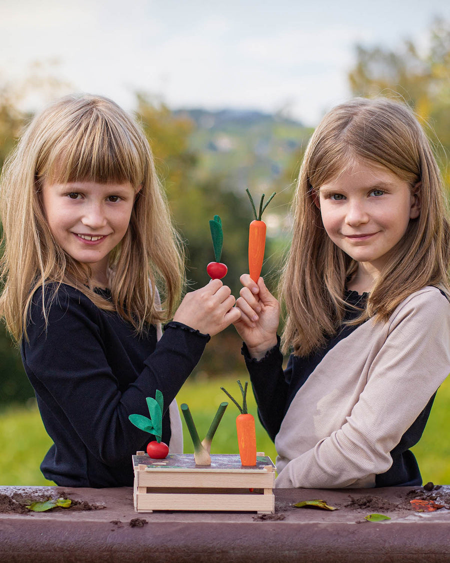 Two children playing with the Erzi Vegetable Patch Wooden Play Food Set. The child on the left is holding the red radish