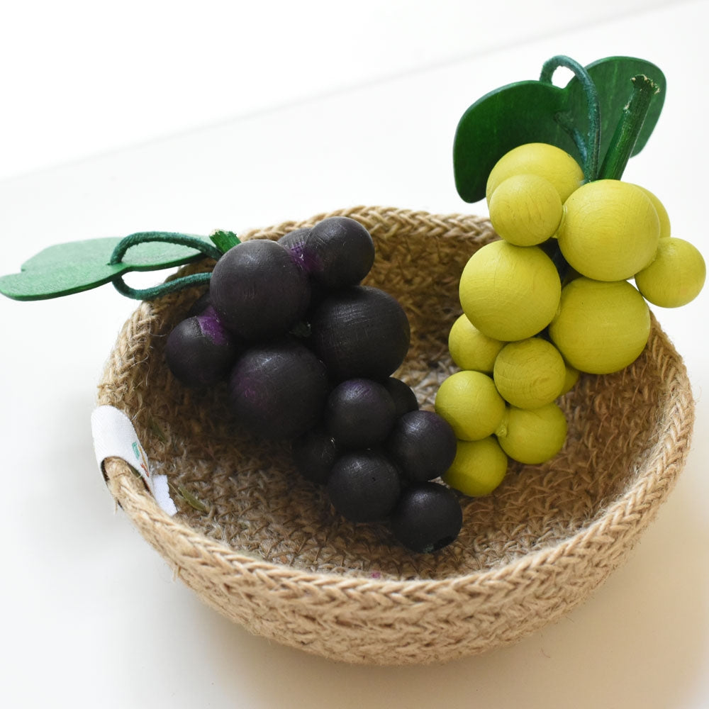 A natural woven bowl holding a bunch of green and purple Erzi wooden grapes.