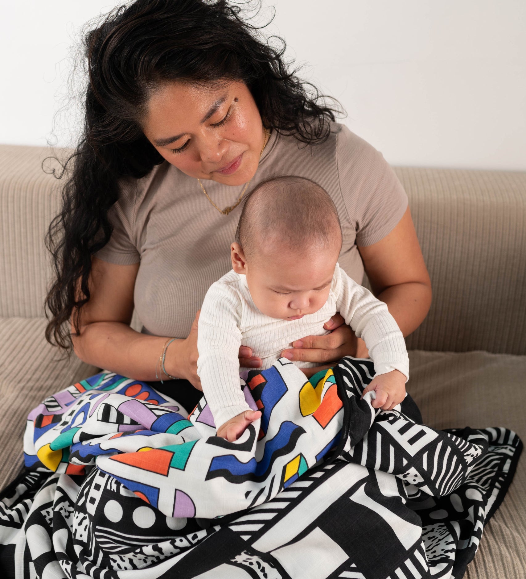 A parent and baby sitting on a sofa, they have a Etta Loves x Camille Walala Muslin Blanket covering their knees. 