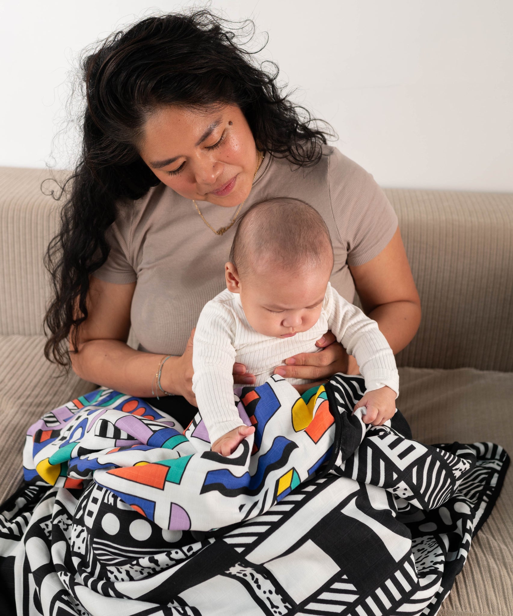 A parent and baby sitting on a sofa, they have a Etta Loves x Camille Walala Muslin Blanket covering their knees. 