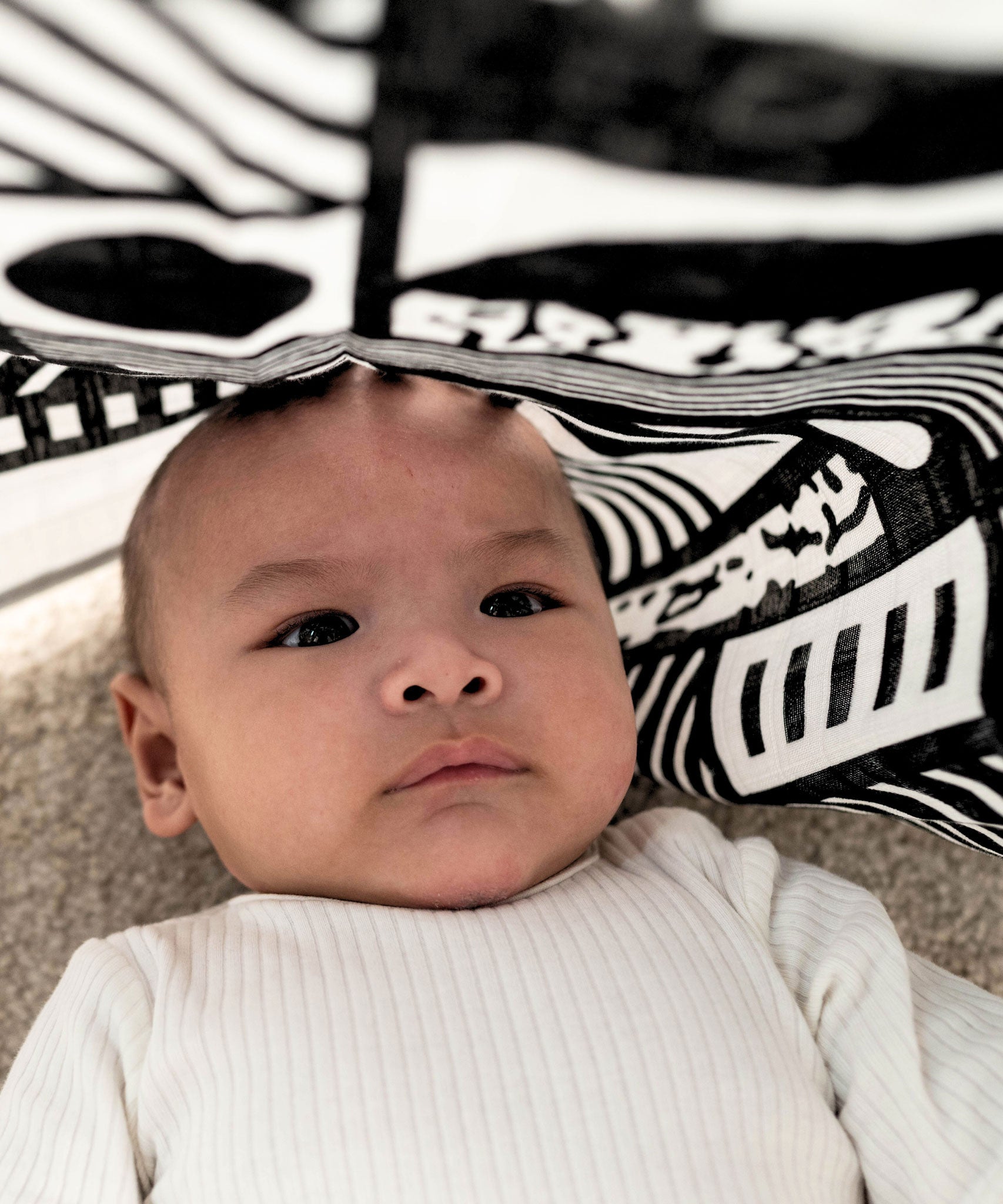 A baby laying underneath a Etta Loves x Camille Walala XL Muslin Square - Mono