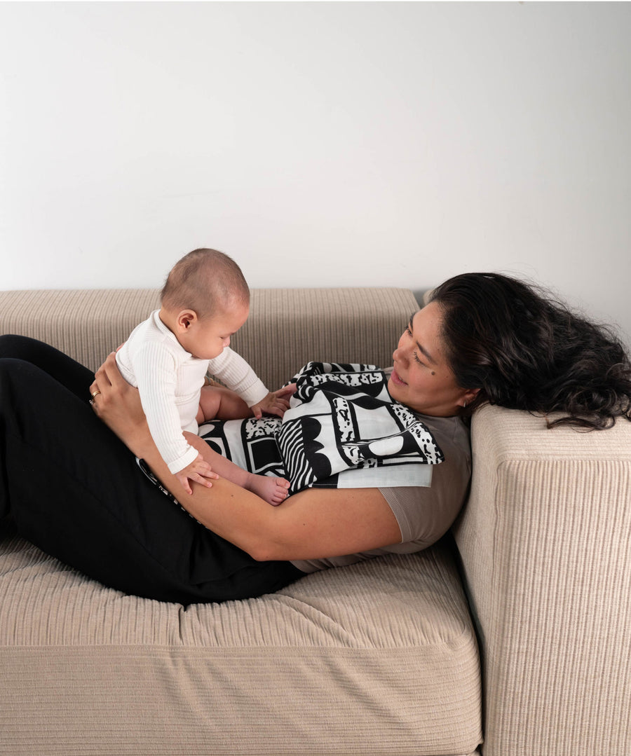 A parent and baby laying on a sofa, the baby is seen looking at the Etta Loves x Camille Walala XL Muslin Square - Mono which has been folded and placed on the parent's chest.