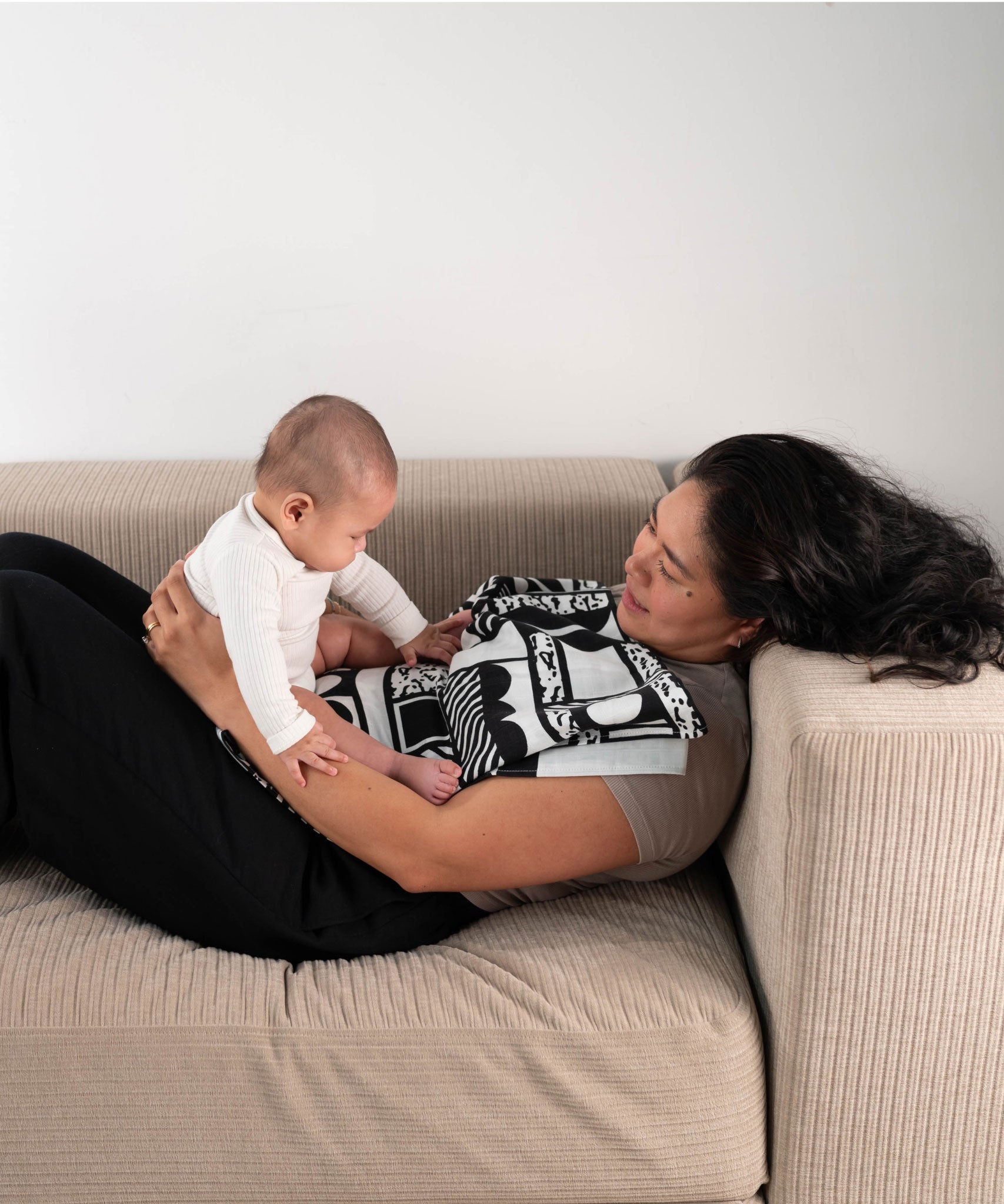 A parent and baby laying on a sofa, the baby is seen looking at the Etta Loves x Camille Walala XL Muslin Square - Mono which has been folded and placed on the parent's chest.