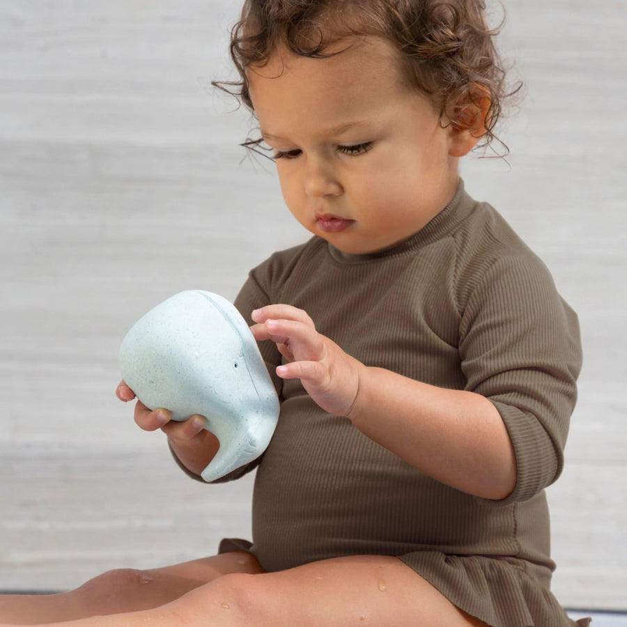 Young child with brown long sleeved dress and brown curly hair is playing with Hevea Squeeze'n'splash Whale Bath toy in Blizzard Blue. Looks very focussed