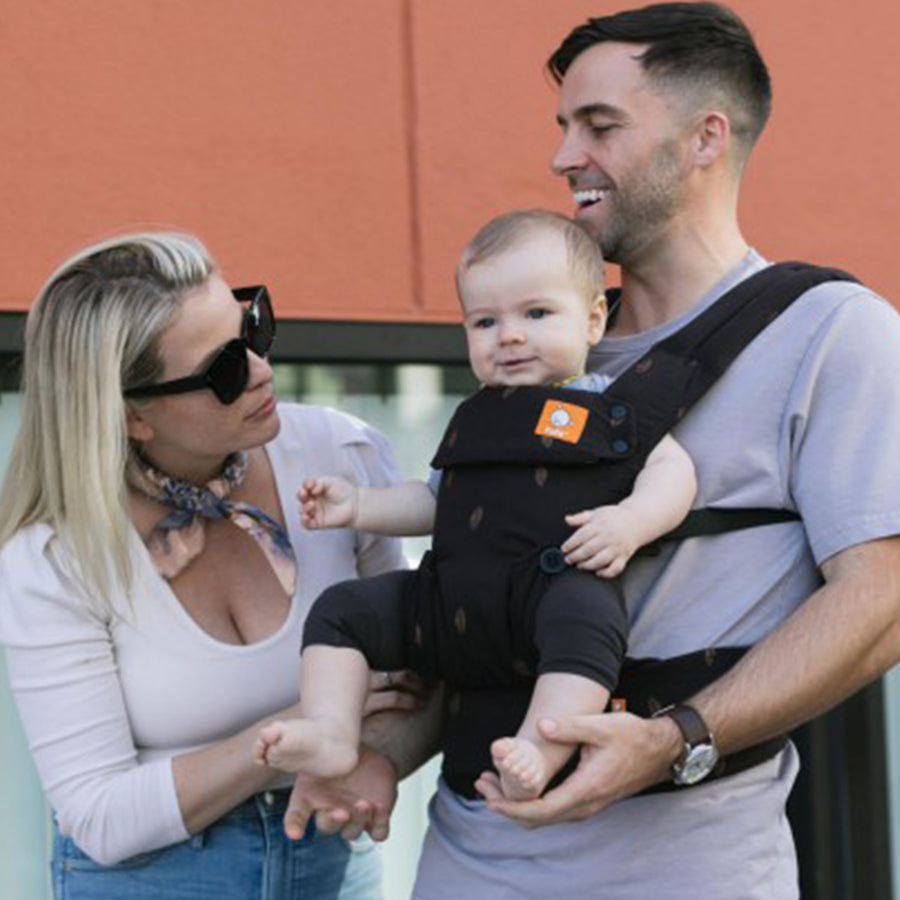 Tula - A man carries a child on his chest wearing a Silva - Explore Baby Carrier. A woman interacts with the child as they walk along.