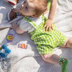 A baby laying on their front wearing the Frugi Alfred Dungaree Outfit - 
 a Macaw green coloured Gingham with Carrots design