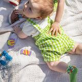 A baby laying on their front wearing the Frugi Alfred Dungaree Outfit - 
 a Macaw green coloured Gingham with Carrots design