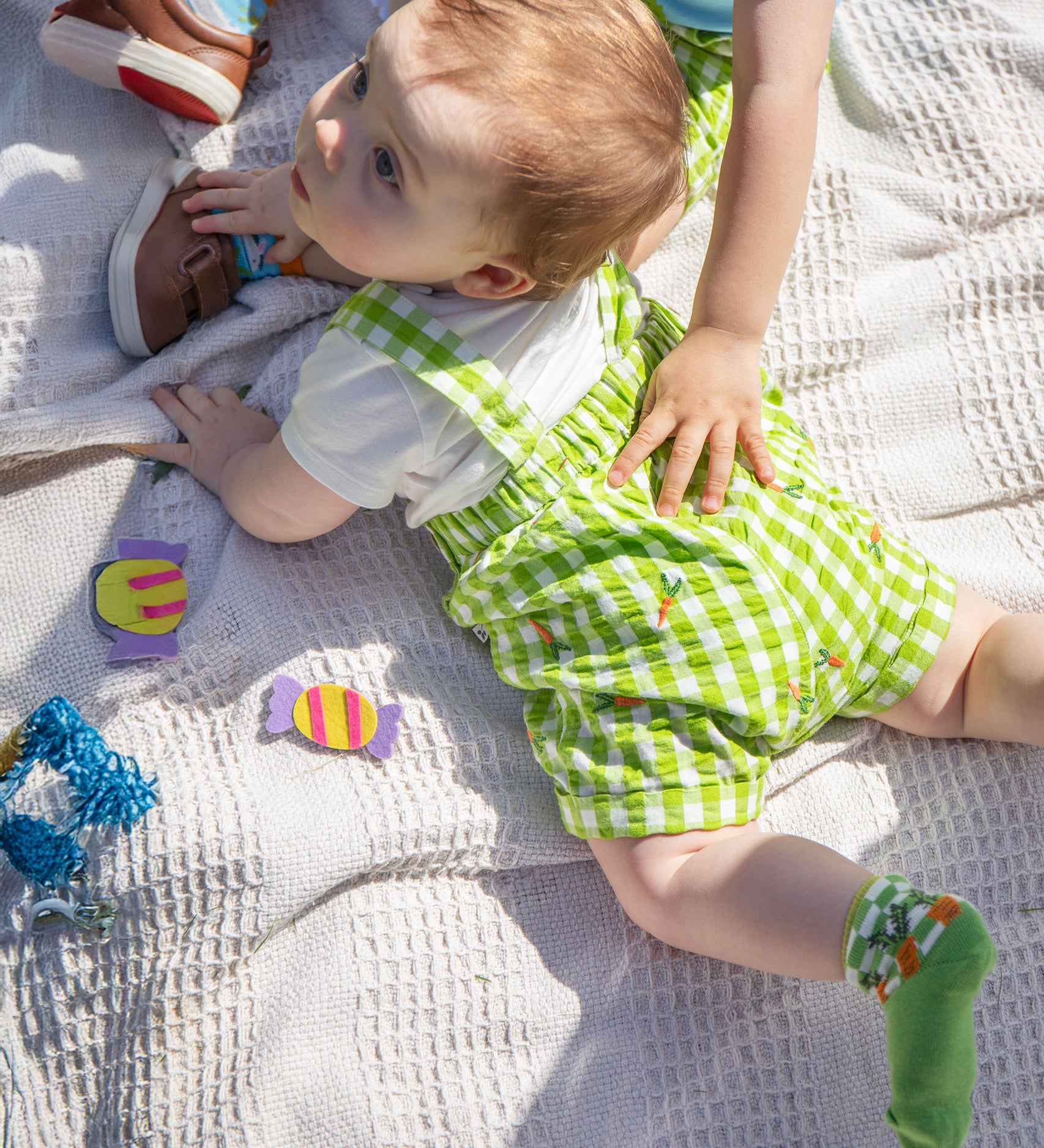 A baby laying on their front wearing the Frugi Alfred Dungaree Outfit - 
 a Macaw green coloured Gingham with Carrots design