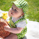 A baby sitting up wearing the Frugi Alfred Dungaree Outfit - 
 a Macaw green coloured Gingham with Carrots design with a coordinating green gingham legionnaires hat