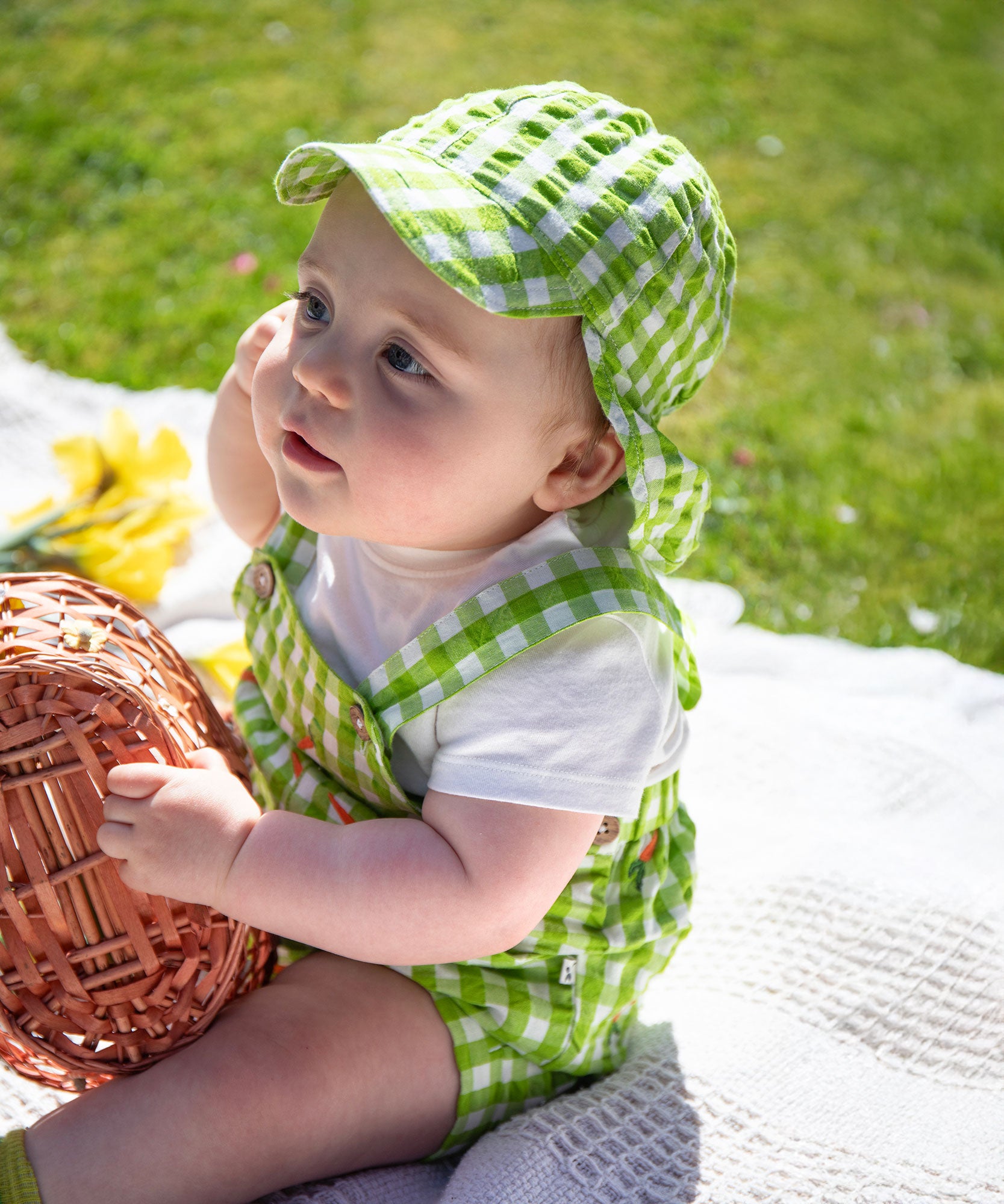 A baby sitting up wearing the Frugi Alfred Dungaree Outfit - 
 a Macaw green coloured Gingham with Carrots design with a coordinating green gingham legionnaires hat