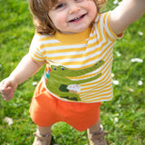 A child wearing the Frugi Crocodile wrap around outfit which includes a yellow striped t-short with a crocodile applique detail and orange coloured shorts.